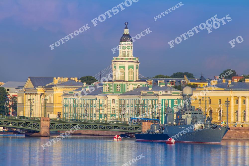 На фотографии изображена Кунсткамера, одно из самых известных зданий Санкт-Петербурга, освещенное мягким утренним светом. На переднем плане стоит военный корабль, пришвартованный у берега реки Невы. Величественная архитектура Васильевского острова гармонично сочетается с отражениями в воде, создавая уникальный городской пейзаж.